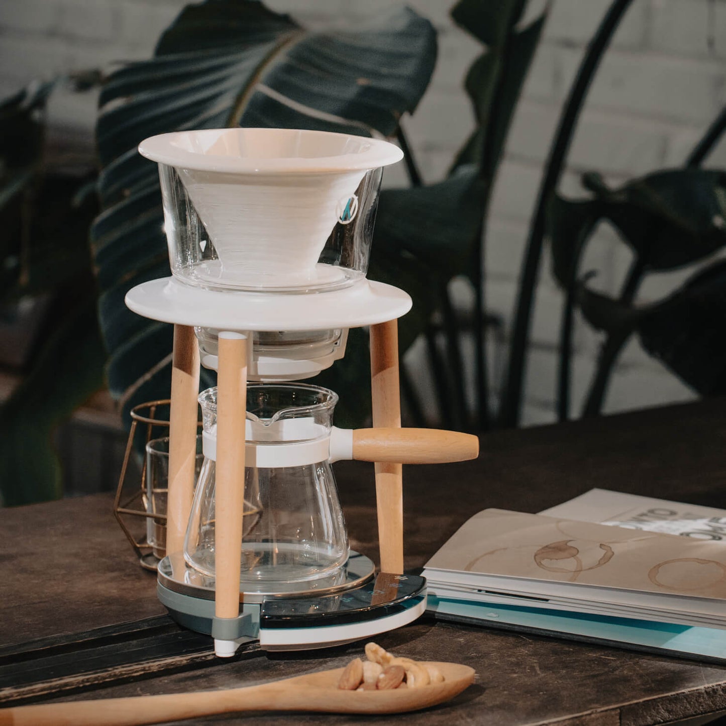 A Wabilogic™ SENZ V™ Pour-Over Smart Coffee Maker set on a dark wooden table, featuring a white cone-shaped filter in a clear glass dripper, supported by a white and light wood stand, with a glass carafe below. A wooden spoon with coffee beans sits in the foreground, and a book is on the right, with large green leaves and a white brick wall in the background.