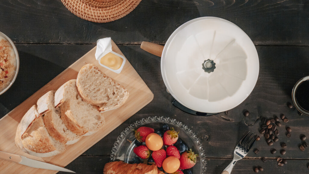 Wabilogic™ SENZ V™ Pour-Over Smart Coffee Maker with a wooden handle sits on a dark rustic wooden table surrounded by breakfast food props including sliced bread on a wooden board, fresh strawberries, coffee beans, and a cup of coffee.